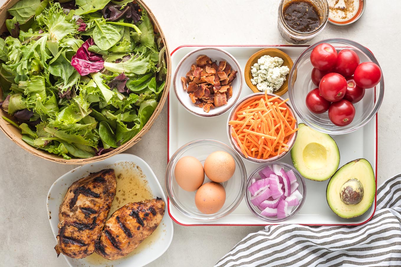 Overhead view of salad greens, grilled chicken, crumbled bacon, eggs in shells, shredded carrots, cheese, cherry tomatoes, red onion, and cut avocado.