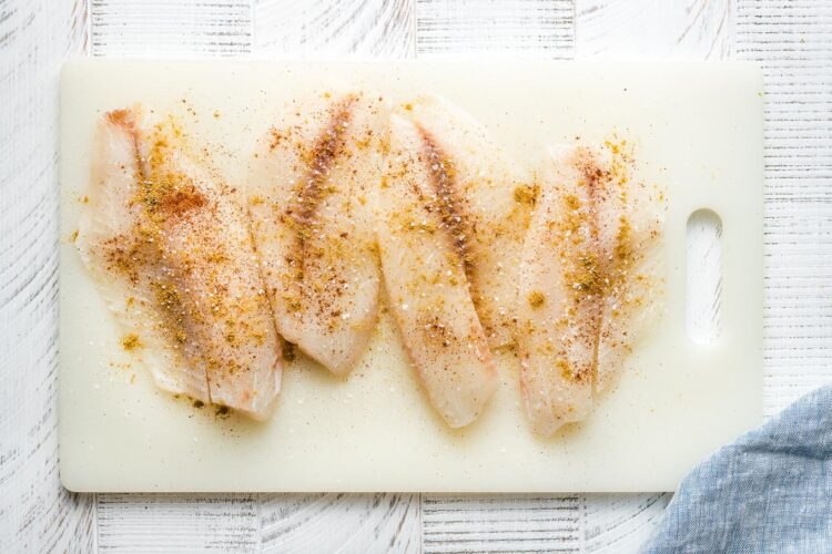 Seasoned tilapia filets on a white cutting board.