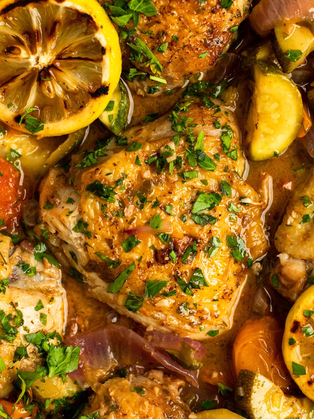 Chicken thighs cooking in a sauce with grilled lemon and veggies.