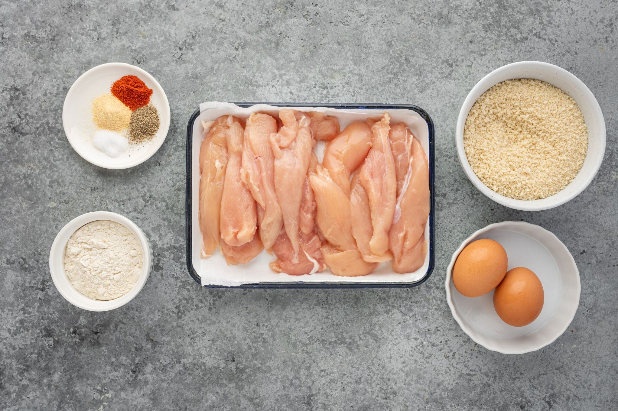 Raw chicken tenders, breadcrumbs, spices, and flour divided into portions.
