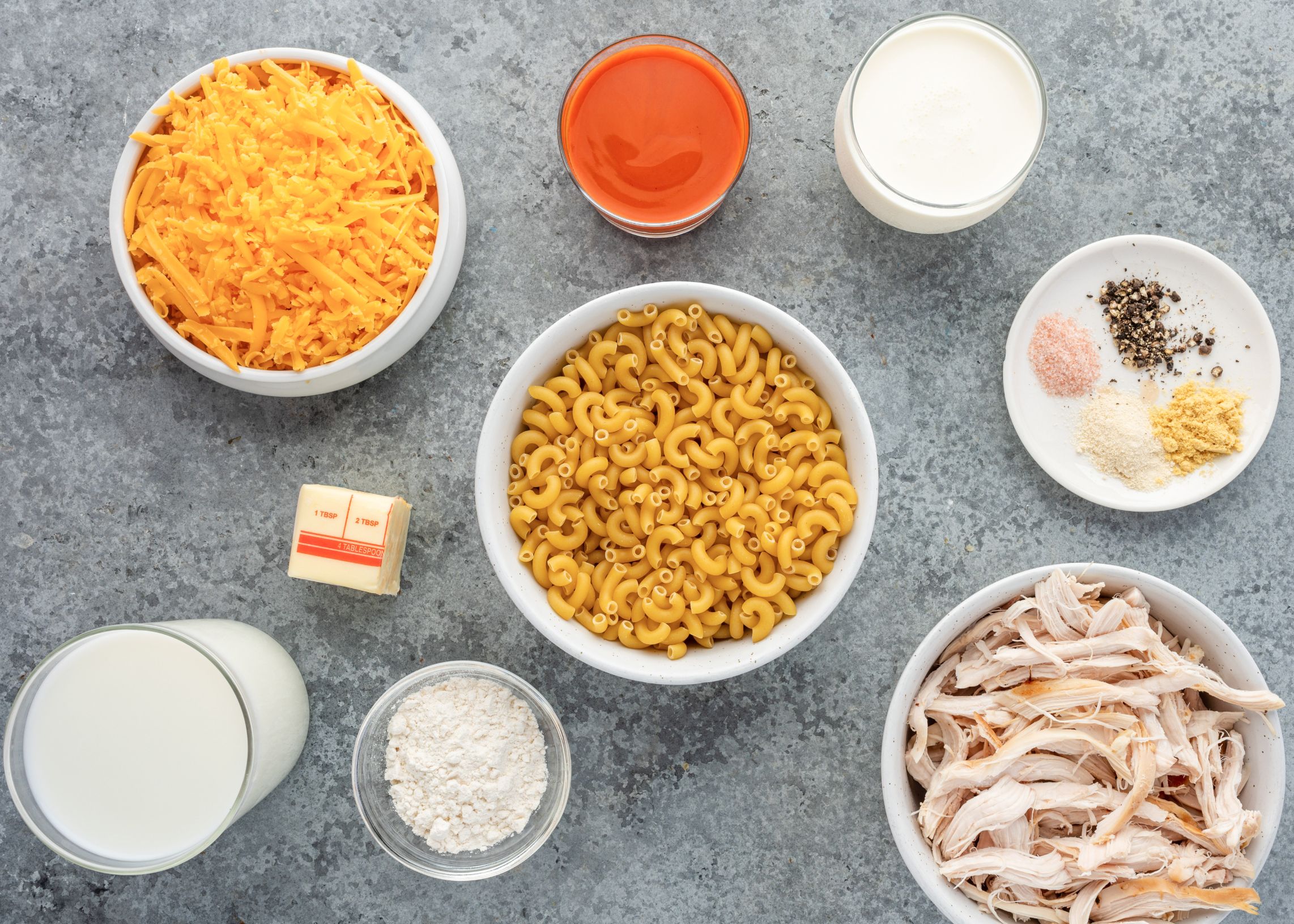 Uncooked noodles, shredded cheese, milk, cream, spices and shredded rotisserie chicken divided into bowls.